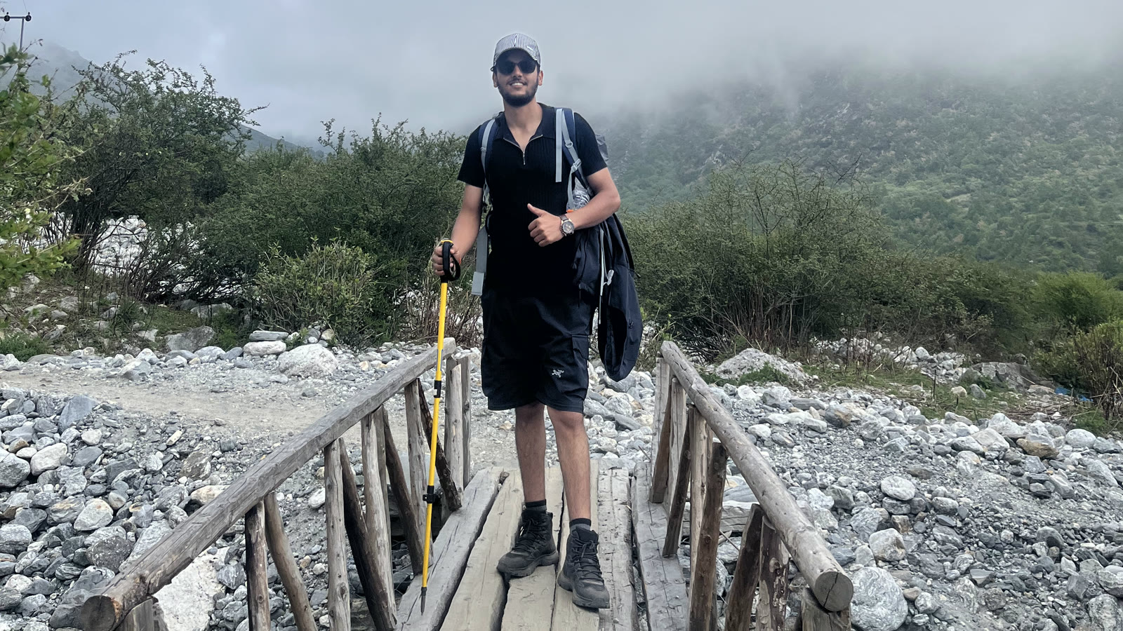 Sam Paudel crossing a log bridge over a boulder riverbed with low cloud sitting on the ridge behind him