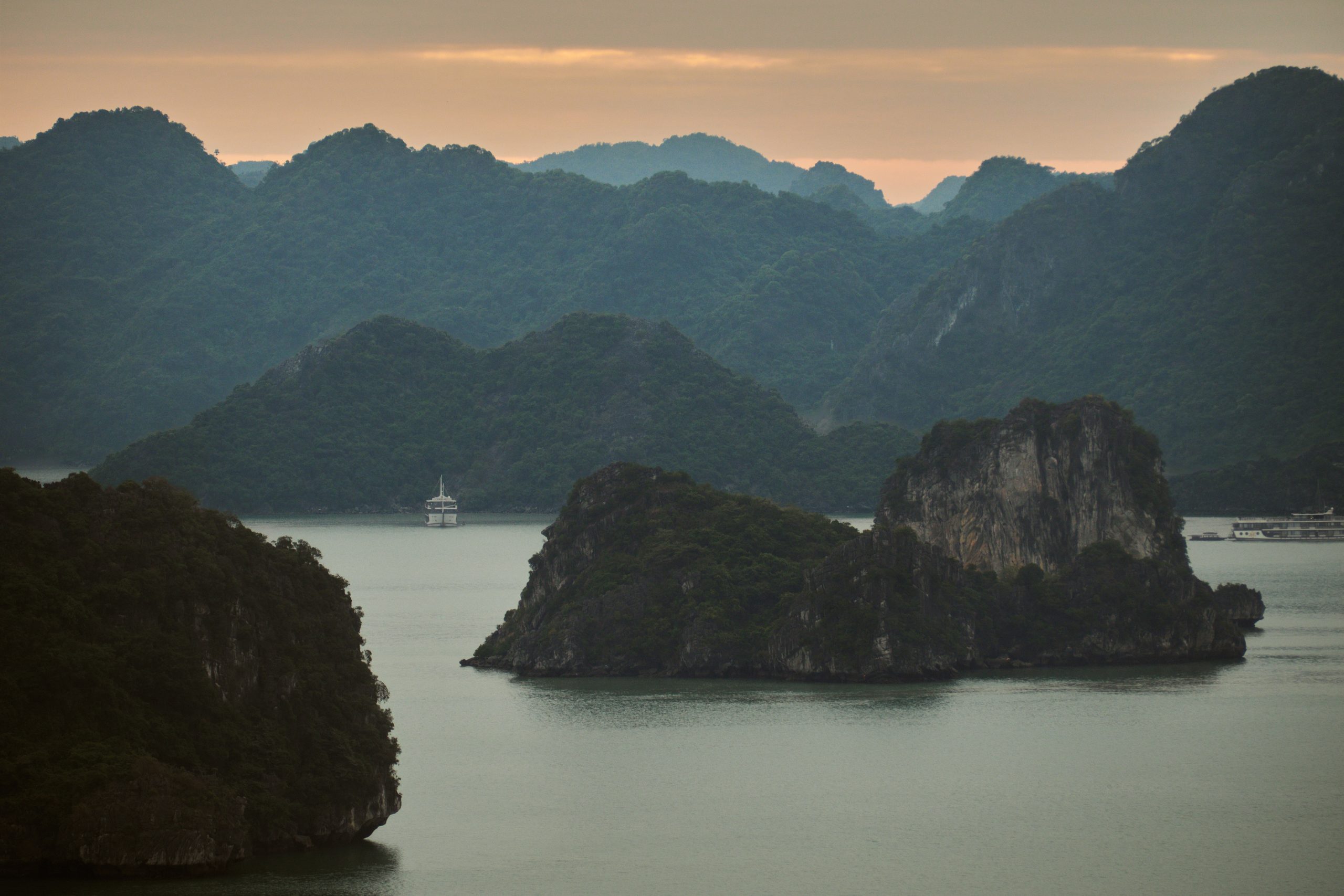 Forested limestone karst islands and a cruise ship on the still water of Ha Long Bay at dawn